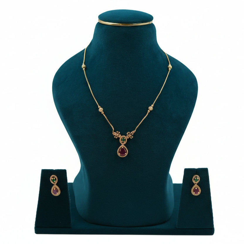 Lalithambike delicate 1-gram gold chain featuring ruby and emerald teardrop pendant with matching earrings on a teal jewelry bust.